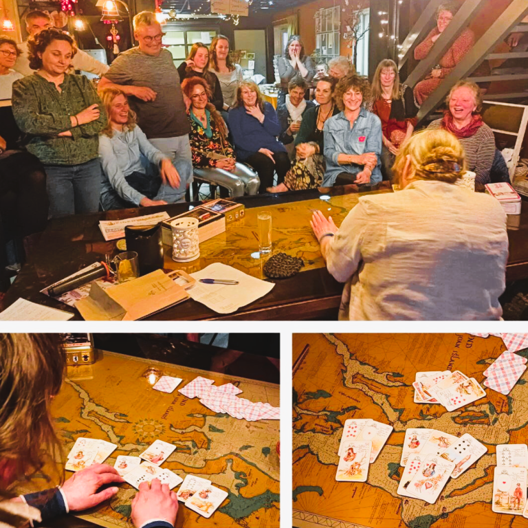 Een vrouw zit aan een grote tafel, omringd door geïnteresseerde deelnemers. Ze legt kaarten uit in een live demonstratie.